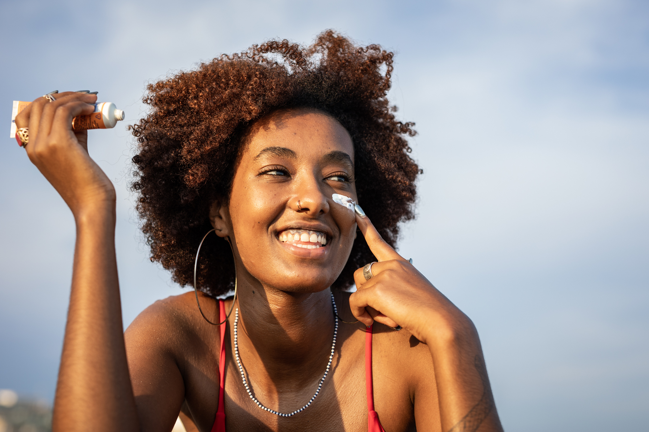 A person with curly hair smiles while applying sunscreen to their nose, wearing a tank top and hoop earrings