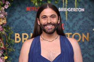 Jonathan Van Ness smiles at a "Bridgerton" event wearing a sleeveless V-neck top and necklace. The background shows floral elements and "Netflix" and "Bridgerton" logos