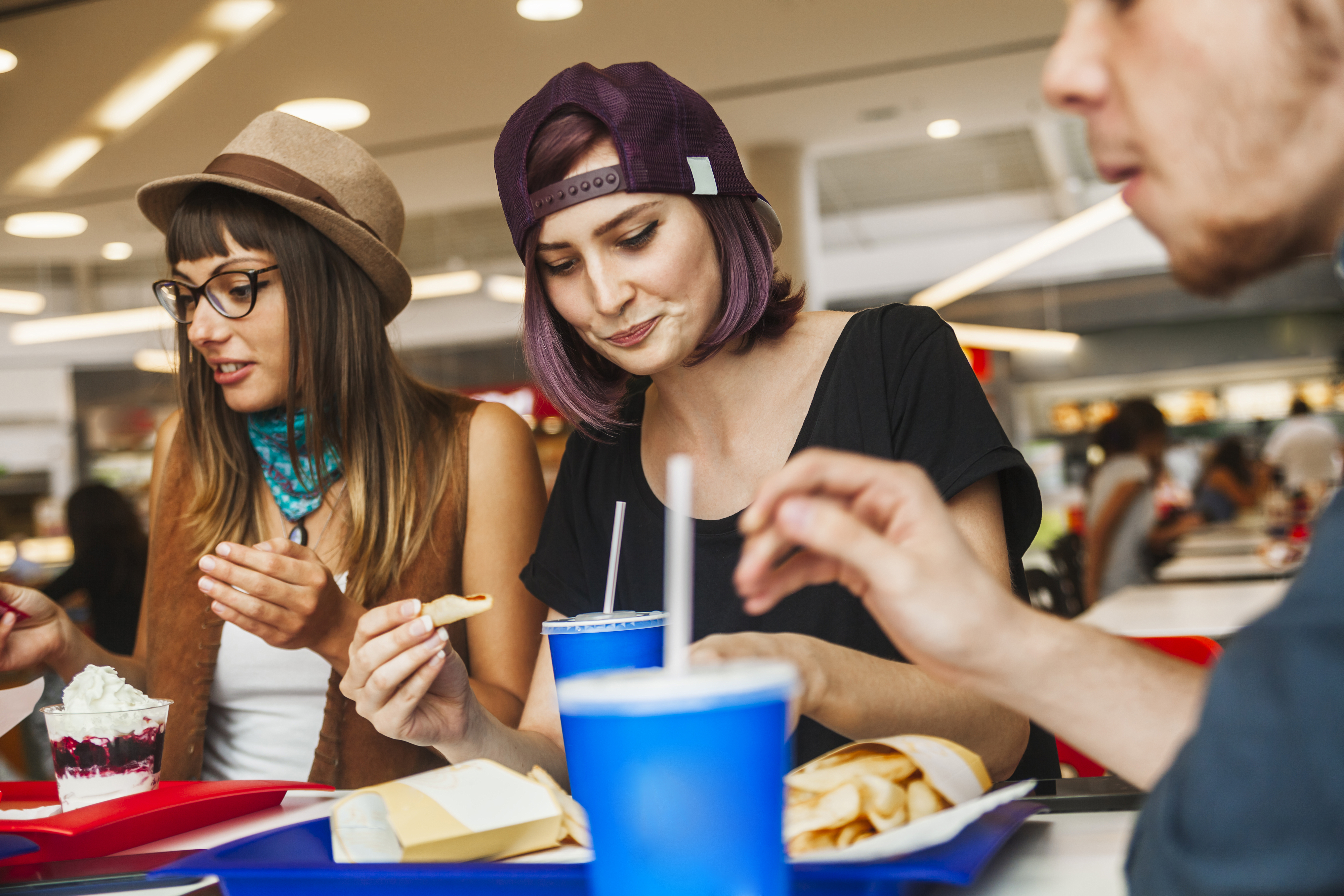 Three people enjoy a meal at a fast-food restaurant in a mall. One person wears glasses and a hat, another wears a backward cap. They are eating fries and drinking soft drinks.