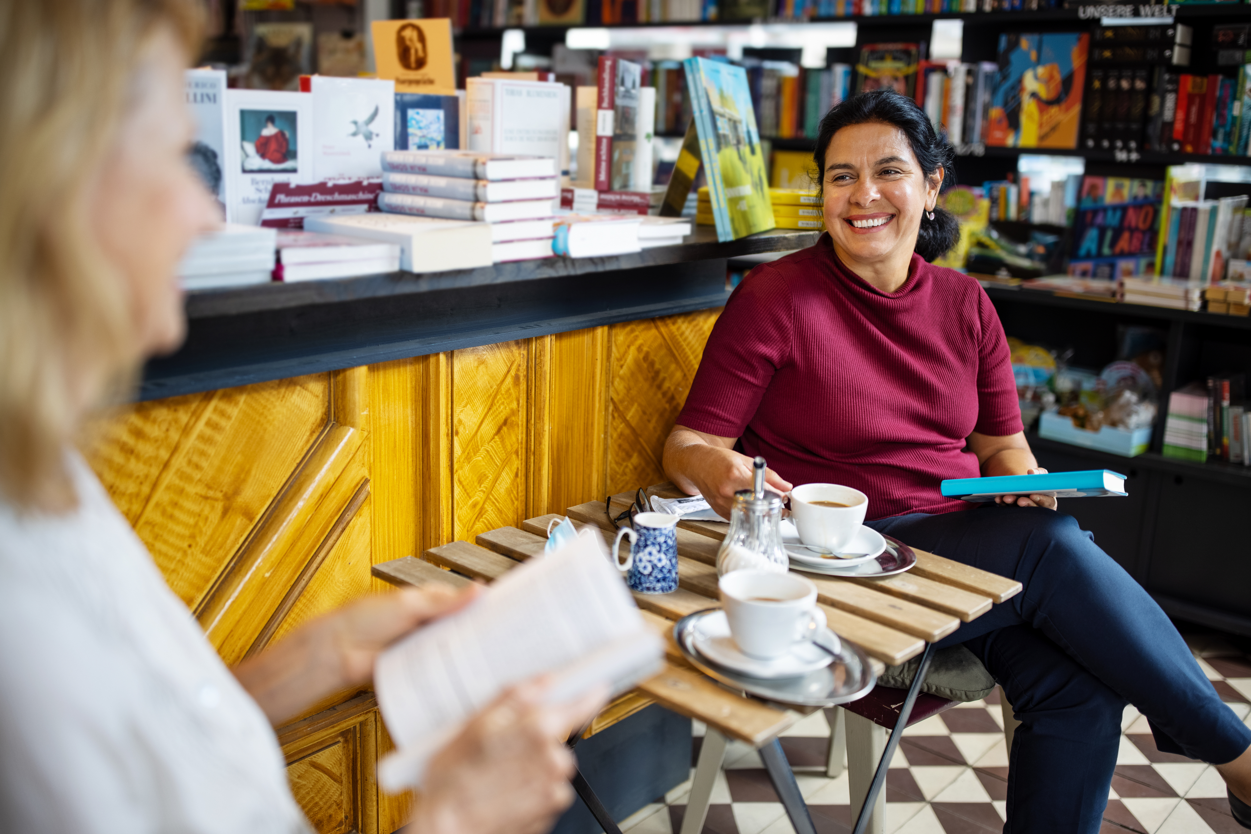 Two women having a conversation over coffee at a table in a bookstore, with one woman holding a book and smiling at the other