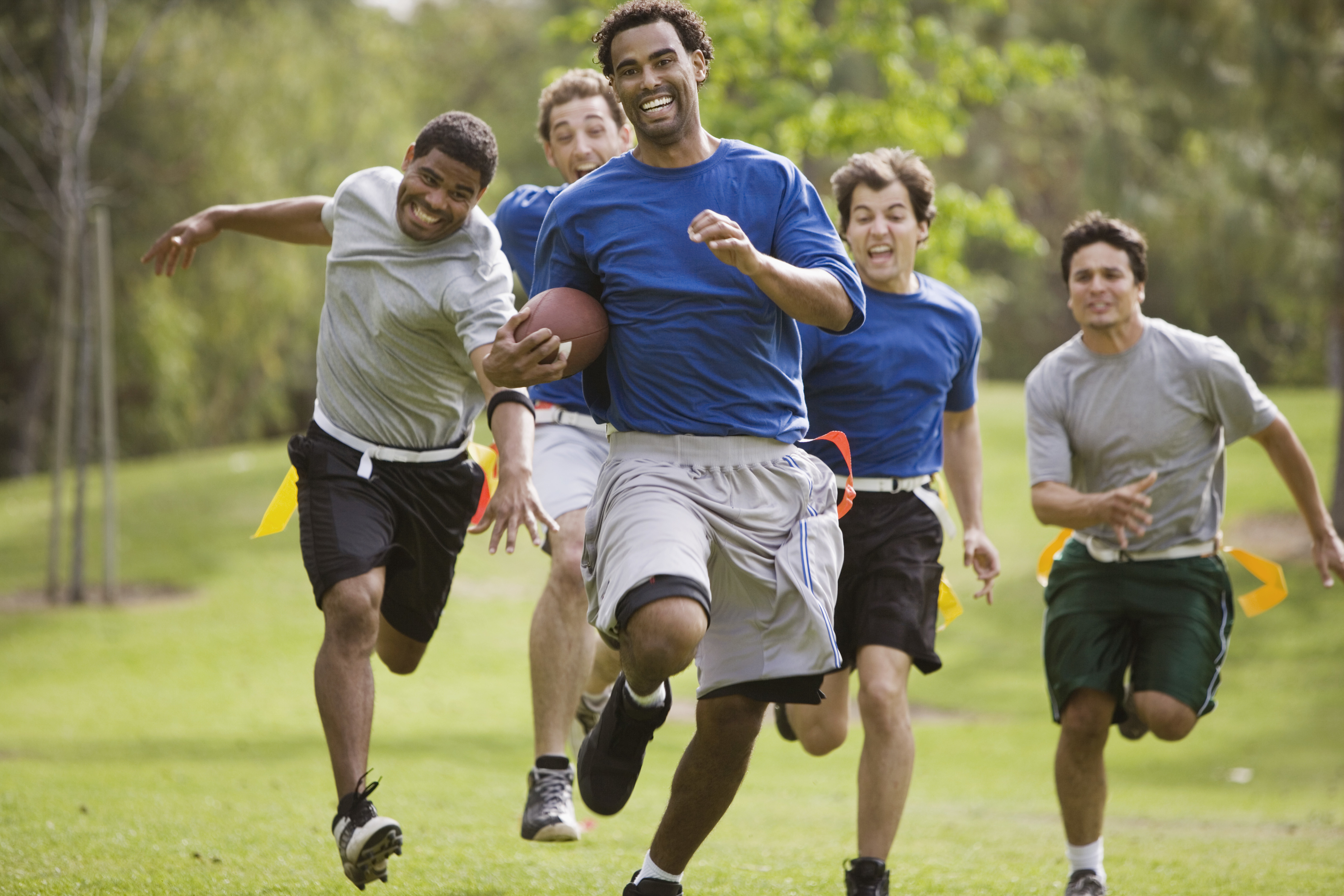 A group of five men playing flag football in a park, with one man holding the football and running while others chase him, all smiling and appearing to have fun