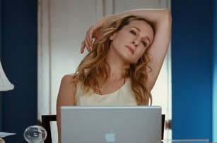 A woman, seated at a desk with an Apple laptop, leans back with one arm resting on top of her head