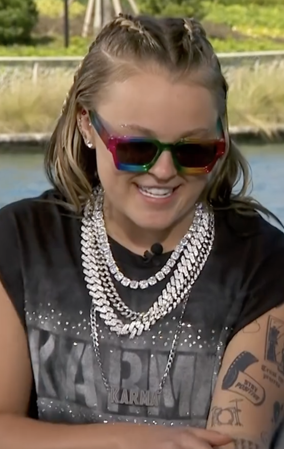 JoJo Siwa smiles while wearing rainbow sunglasses, layered chain necklaces, and a &quot;KARMA&quot; t-shirt. Her arm tattoos are visible