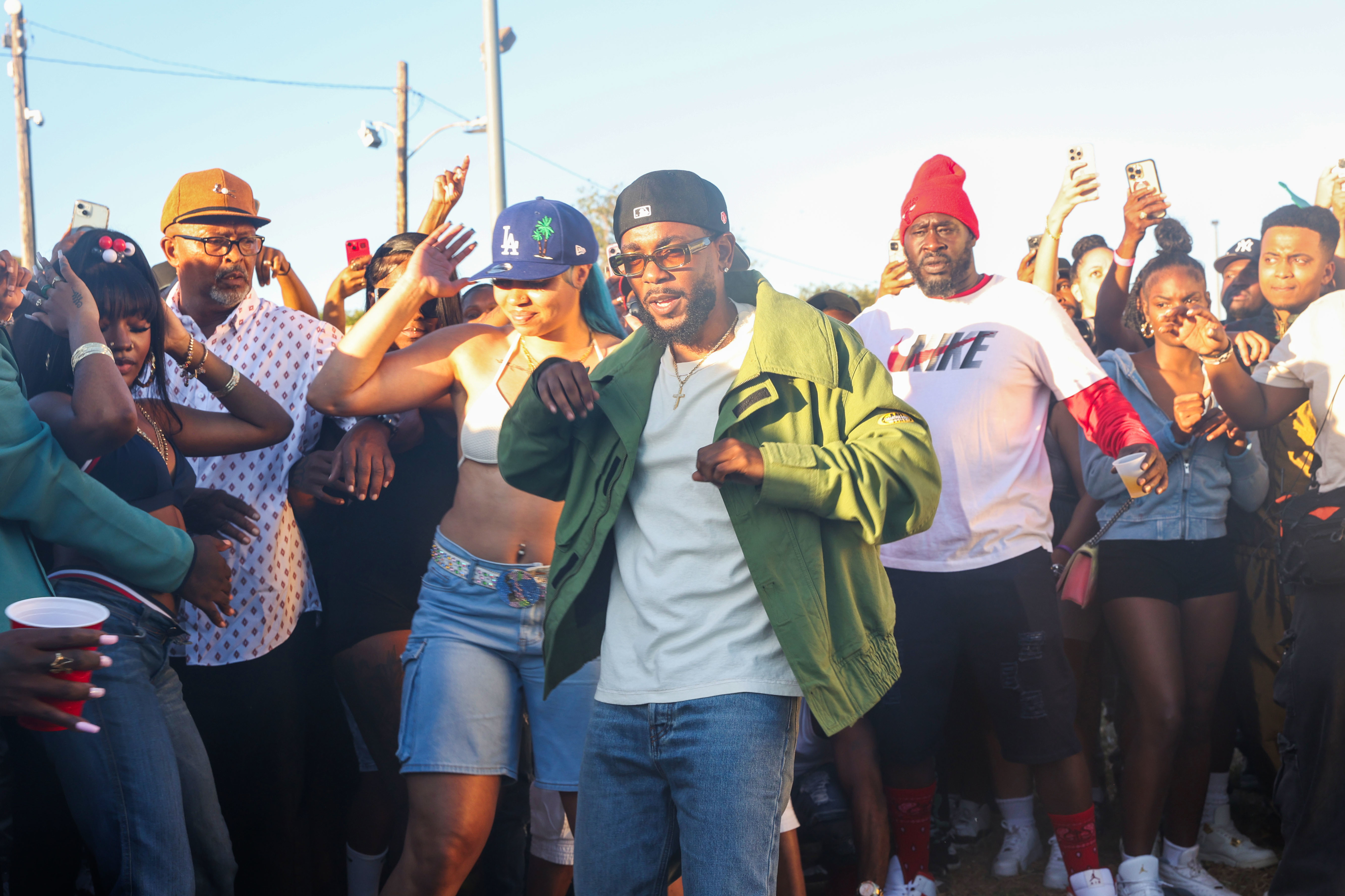 Kendrick Lamar dances in a crowd at an outdoor gathering. Lamar wears a green jacket, white shirt, and black cap. People around him take photos and enjoy the moment