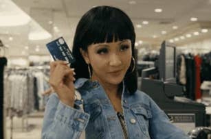 A woman with medium-length dark hair holds up a credit card in a clothing store. She wears hoop earrings and a denim jacket. The store has racks of clothes in the background