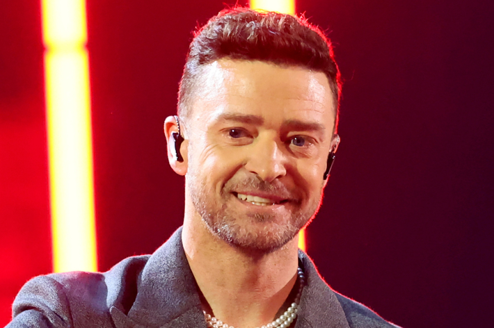 A man with short brown hair wearing a grey blazer and pearl necklace smiles at the camera, standing in front of a stage with bright lights