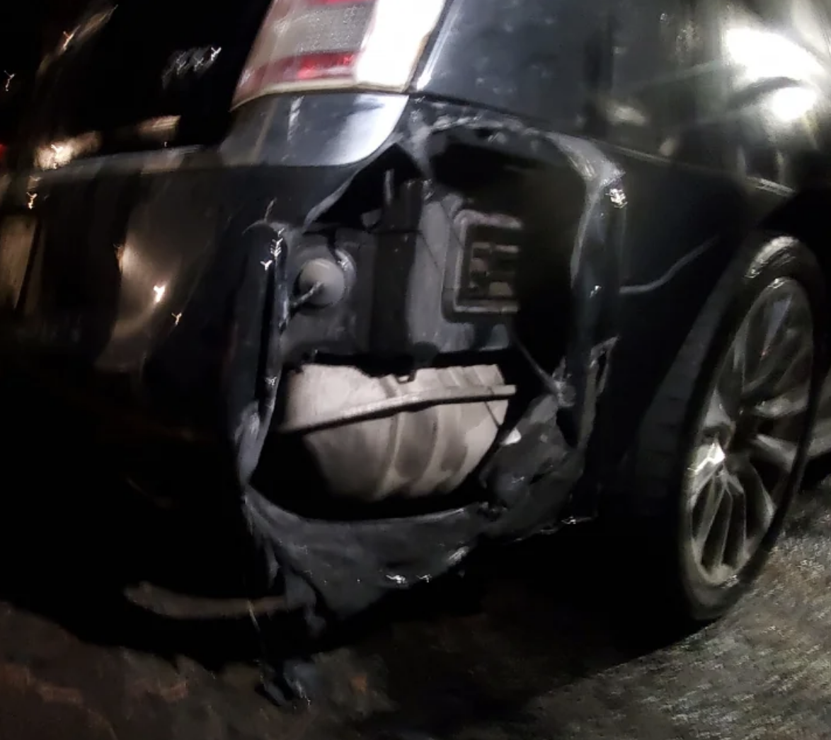 The image shows the back end of a damaged car with a severely dented and torn rear bumper, exposing parts like the exhaust pipe and inner components