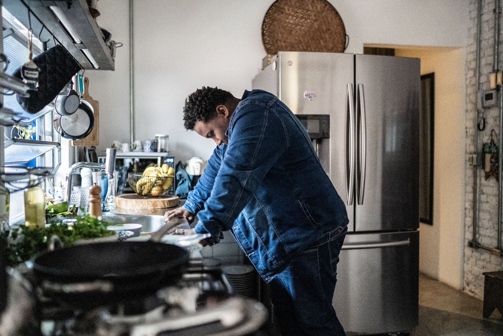 A person stands at a kitchen counter, leaning over a bowl, possibly preparing food. The environment is modern with a stainless steel refrigerator in the background