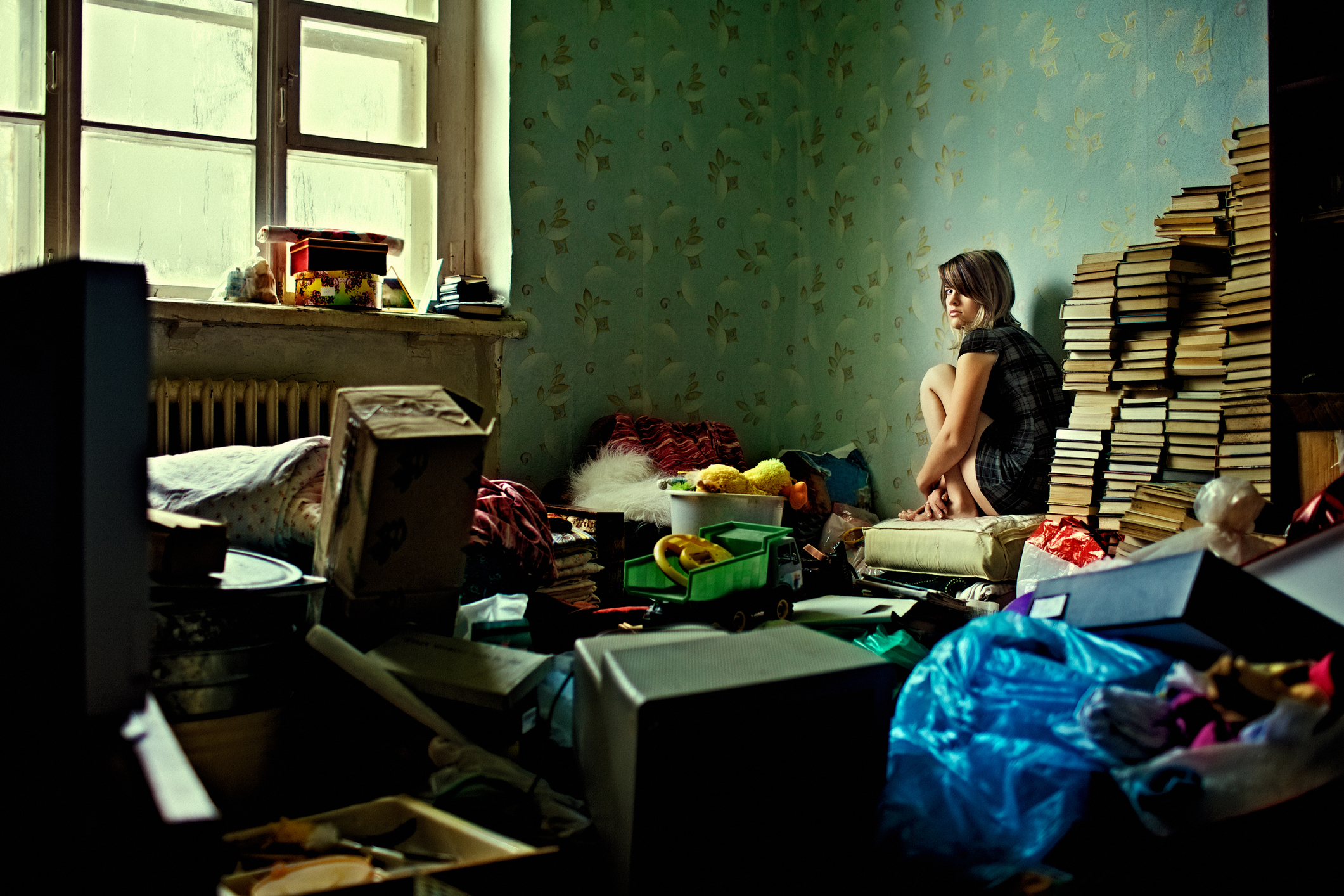 A person is sitting and curled up on a stool in a cluttered, messy room filled with stacks of books, boxes, and various scattered items