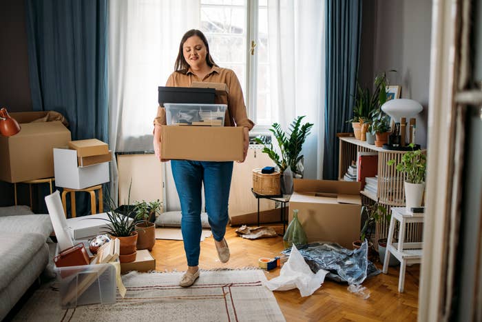 Woman carrying a cardboard box in a room filled with plants, open boxes, and scattered items, appearing to be in the process of organizing or moving