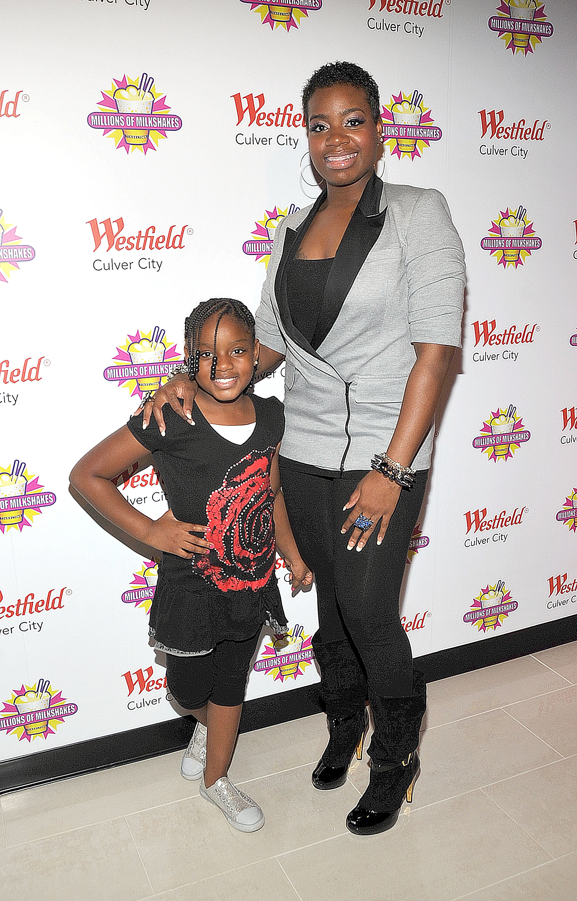 Fantasia Barrino stands with her daughter Zion Quari Barrino at a Millions of Milkshakes event. Fantasia wears a gray blazer and black shirt, Zion wears a black outfit with a rose design