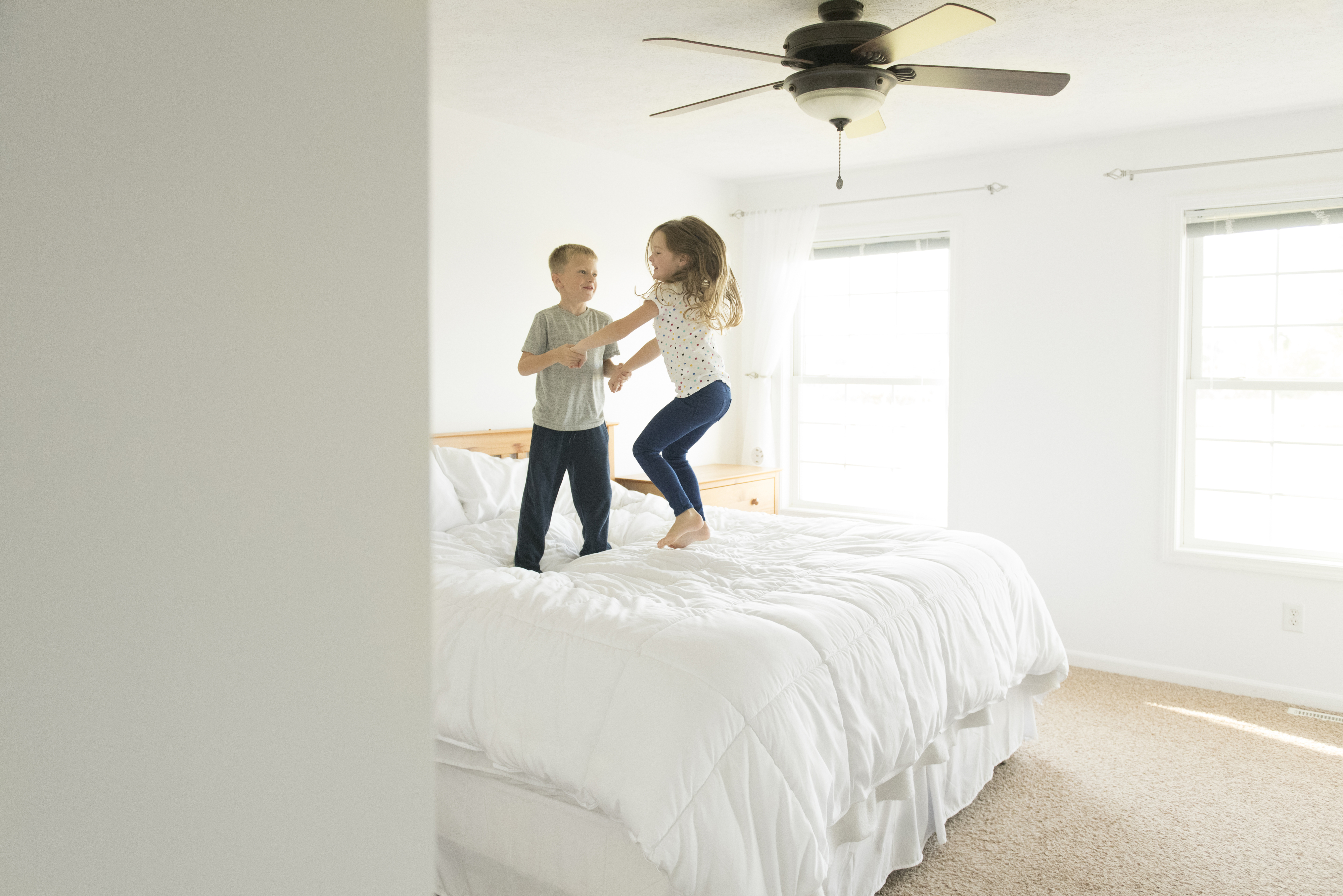 Two children, a boy and a girl, jump on a large white bed in a bright bedroom with a ceiling fan and two windows