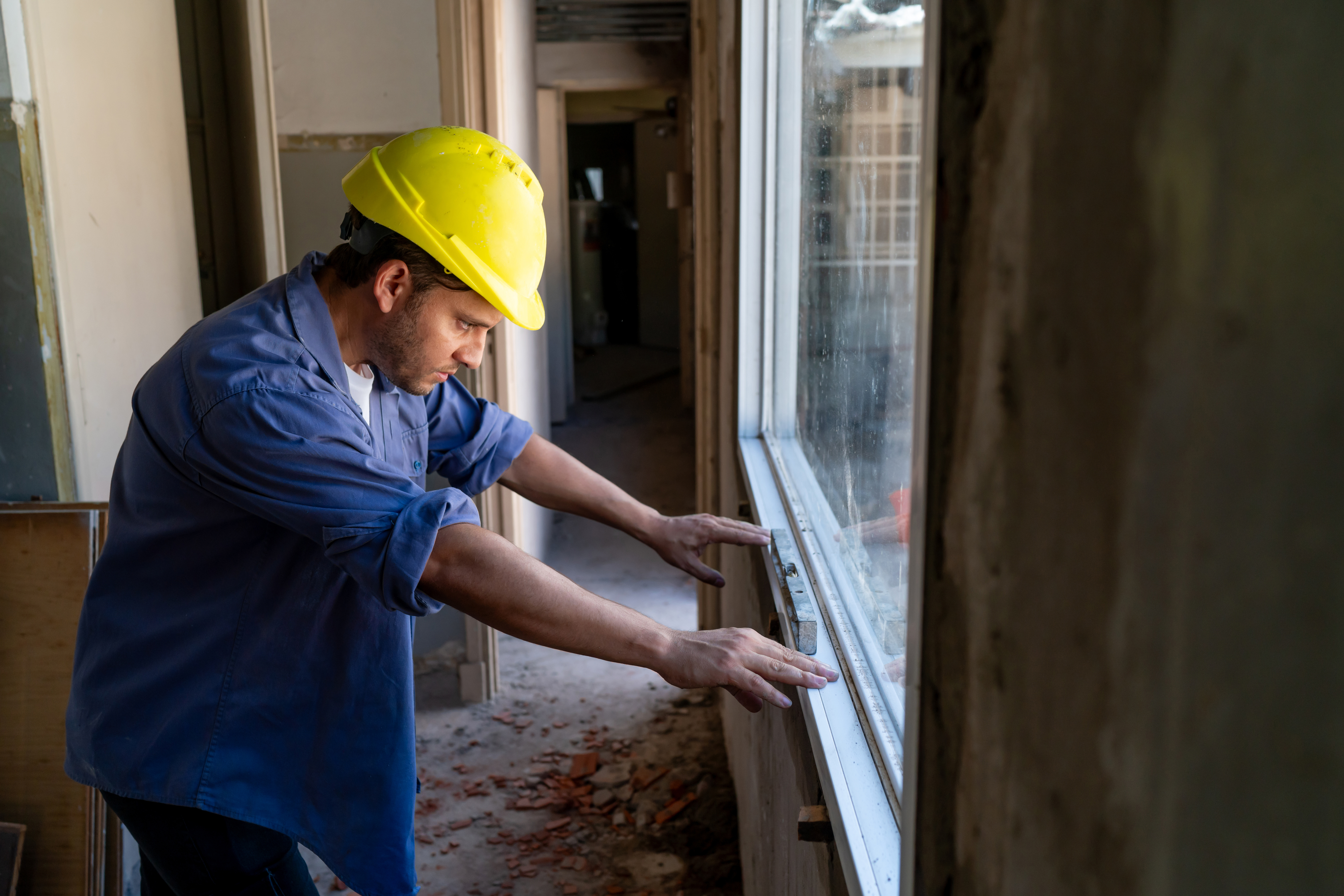 A construction worker wearing a yellow hard hat and blue shirt is adjusting a window frame inside a building