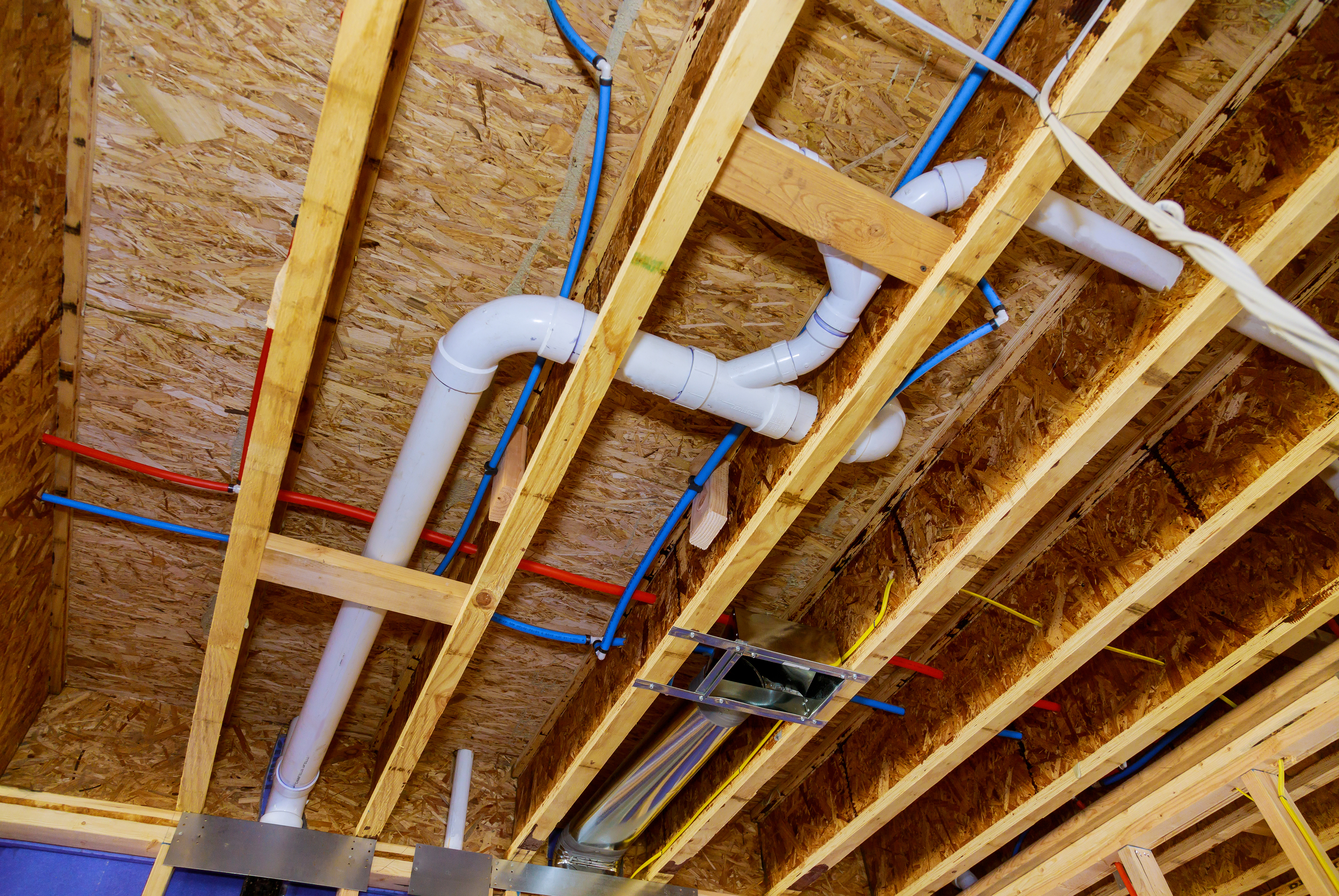 An exposed basement ceiling showing plumbing and electrical wiring installed among wooden beams and panels