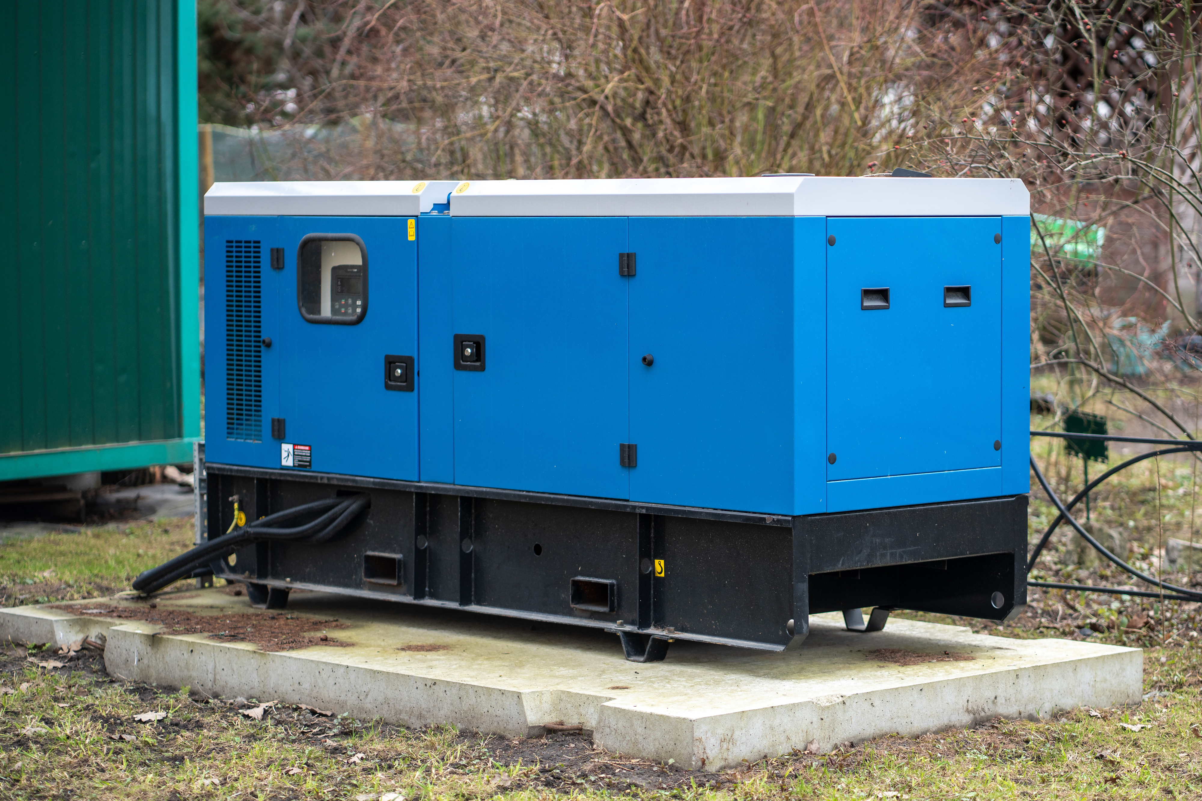 Large blue outdoor generator placed on a concrete slab with several cables connected, surrounded by grass and plants