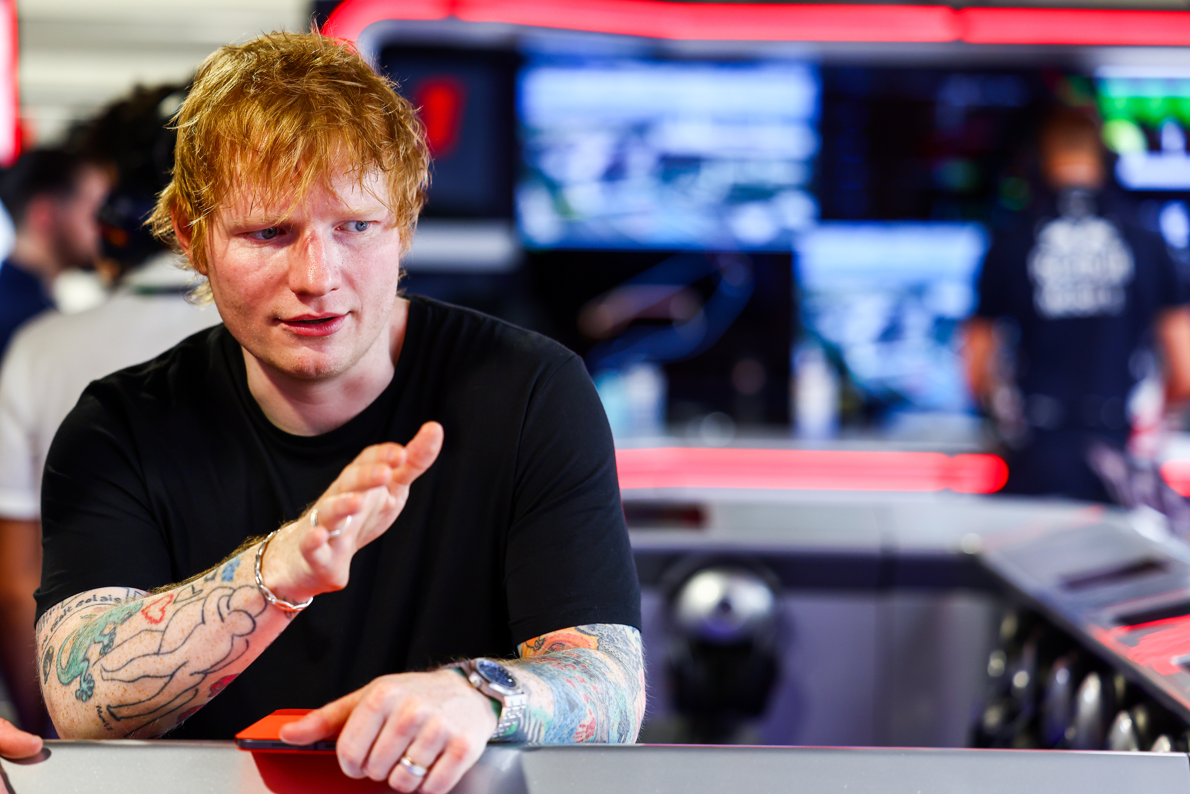 Ed Sheeran gestures while sitting at a console, surrounded by people and screens in the background