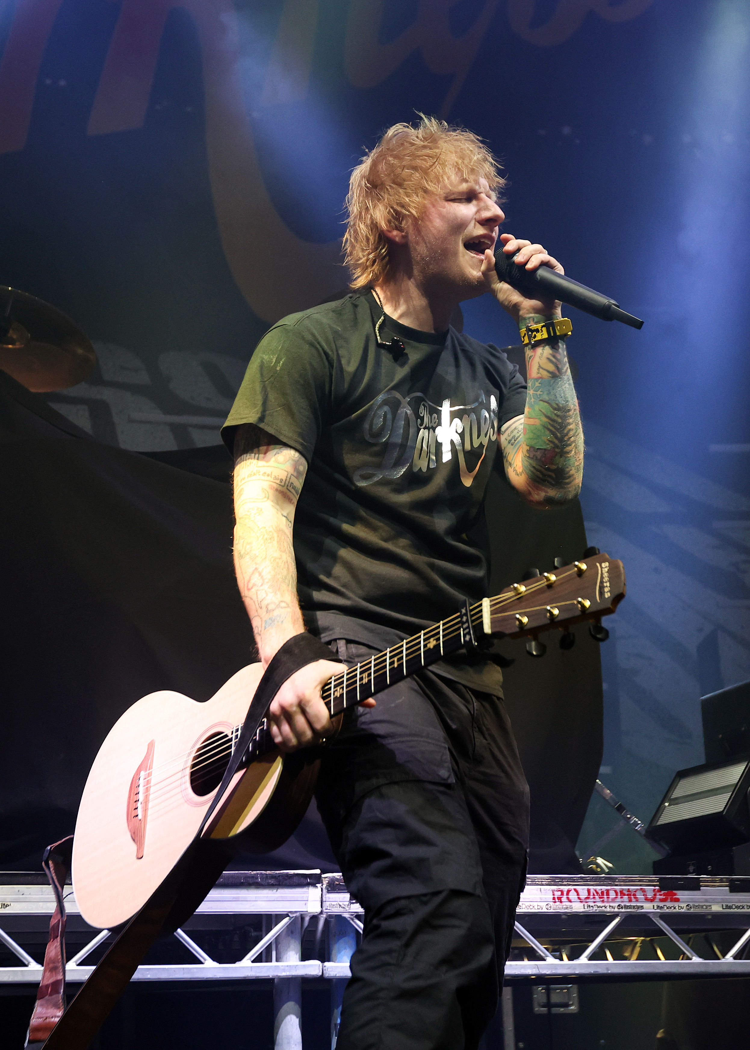 Ed Sheeran performing on stage, holding a guitar and singing into a microphone. He is wearing a casual T-shirt and jeans