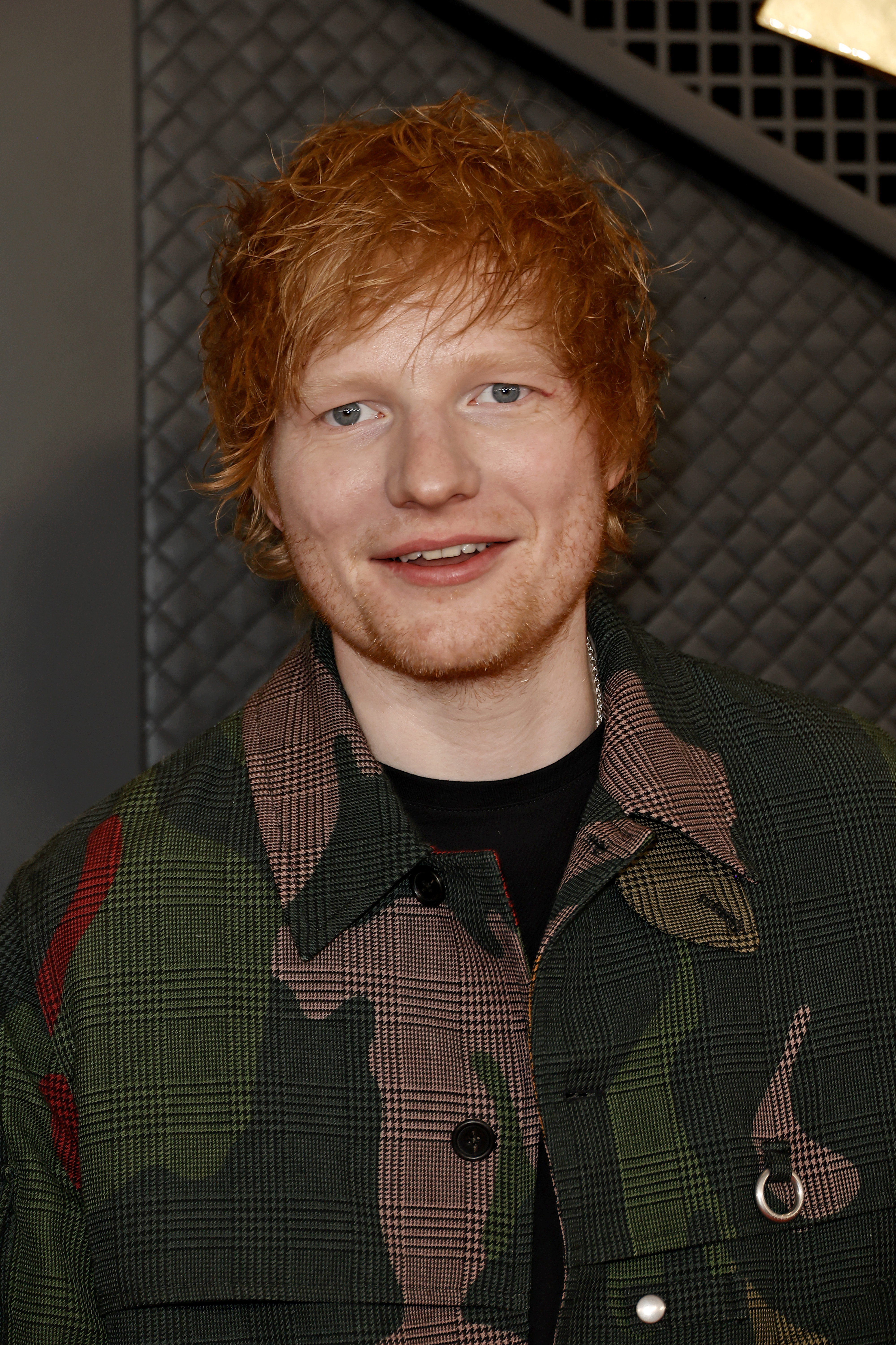 Ed Sheeran smiles, wearing a patterned jacket over a black top. He stands in front of a quilted background