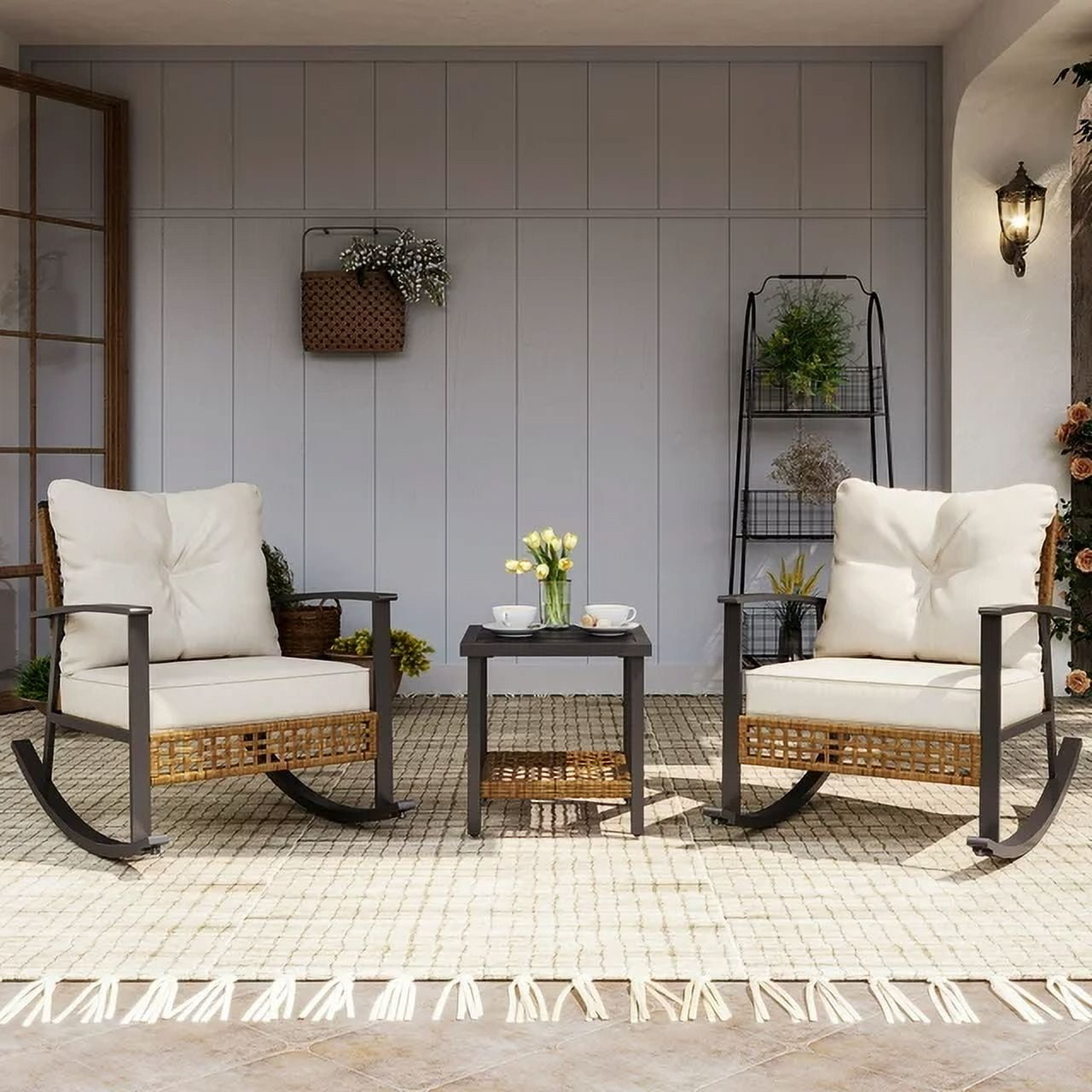 Two cushioned rocking chairs and a small table with a vase of flowers are arranged on a patio. Background includes a shelf with plants and a wall lantern