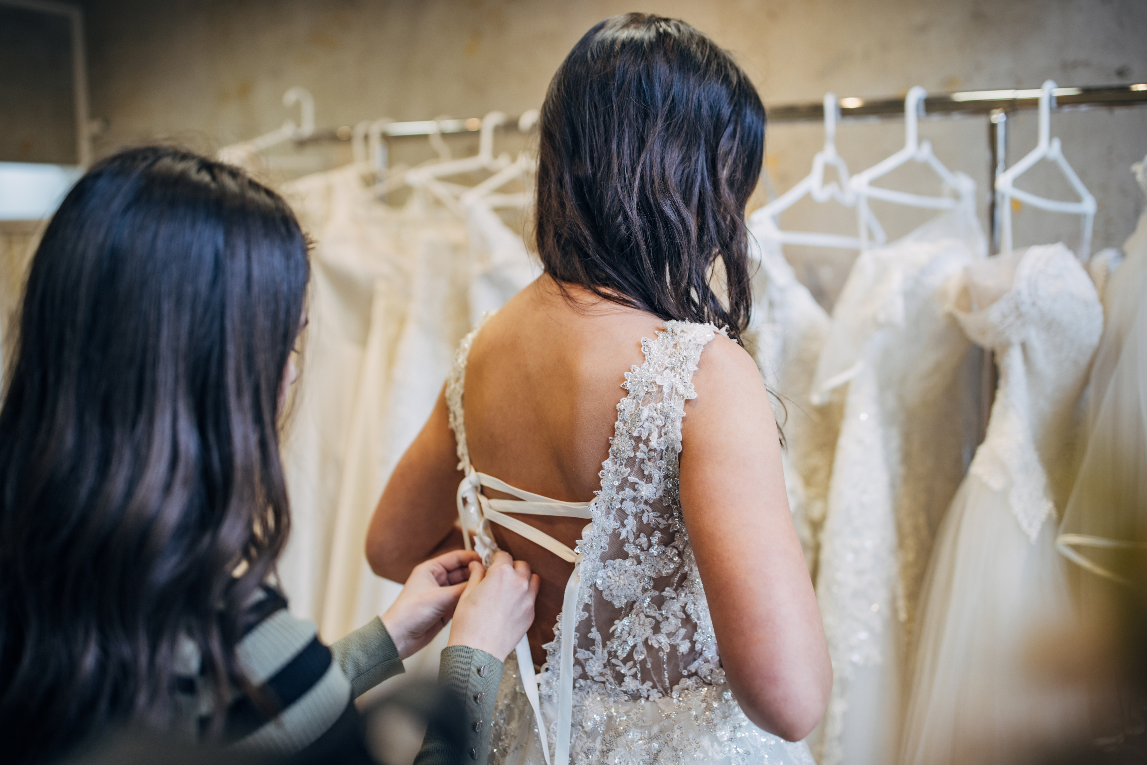 A bride tries on a lace wedding dress. Another person assists with the lace-up back. Various wedding dresses hang on a rack in the background