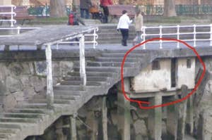 Concrete stairs reaching into the water, with a crumbling section circled in red, highlighting damage and potential hazard. Two people walk on the pier above