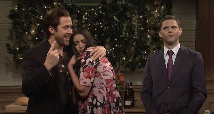 Ryan Gosling in a dark suit points while hugging Cecily Strong in a floral dress; Mikey Day in a suit looks on. Scene from a comedy show skit with warm lighting