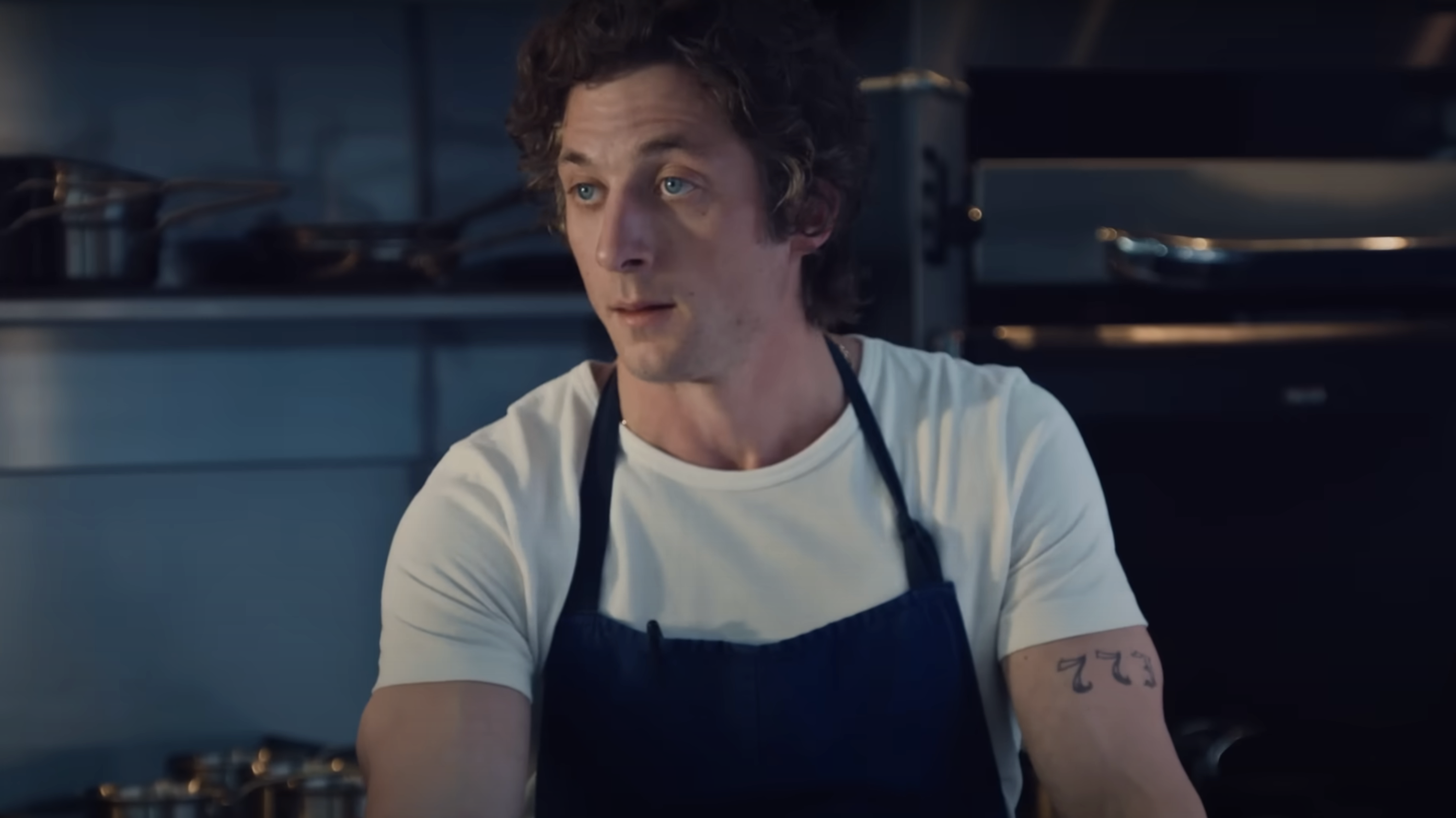 Jeremy Allen White in a  t-shirt and a dark apron stands in a kitchen, with a tattoo of &quot;322&quot; on his left arm