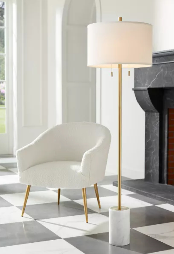 A modern room features a cozy white armchair with gold legs and a stylish floor lamp with a white shade and gold base near a large fireplace. No people are present