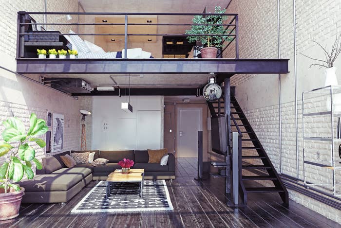 Modern loft apartment with industrial decor, featuring a spacious open living area with a dark sofa, coffee table, plants, and a mezzanine bedroom above