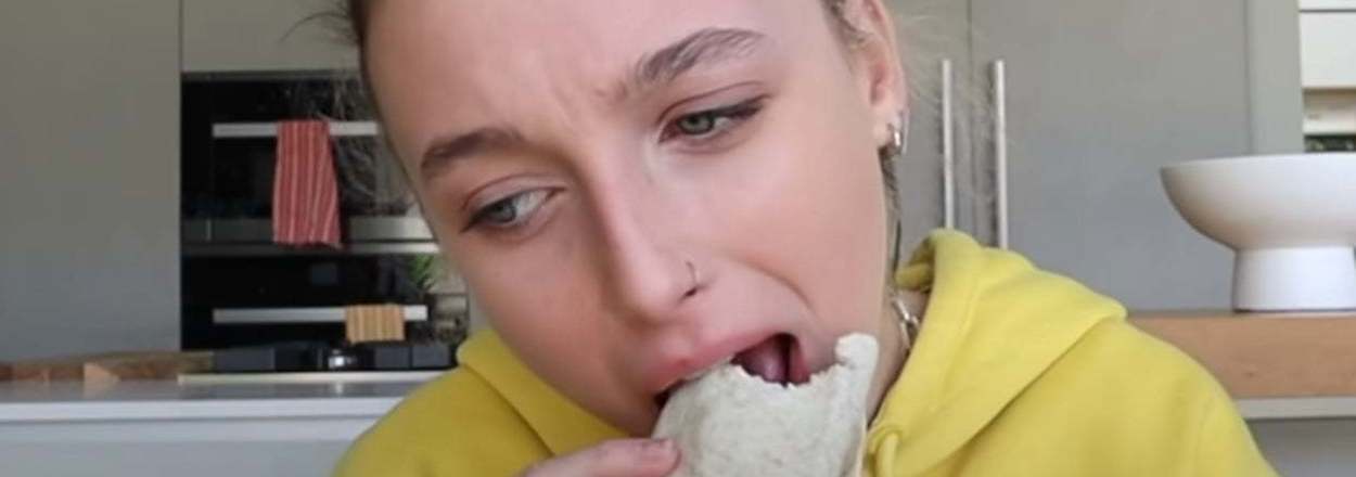 Emma Chamberlain takes a bite of a burrito while seated in a kitchen wearing a hoodie