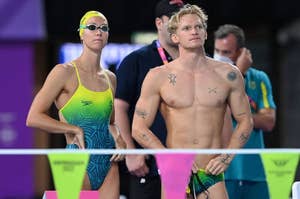 Emma McKeon and Cody Simpson stand poolside in swimwear before a swim event. Emma wears a swim cap and goggles. Cody has tattoos and holds his goggles