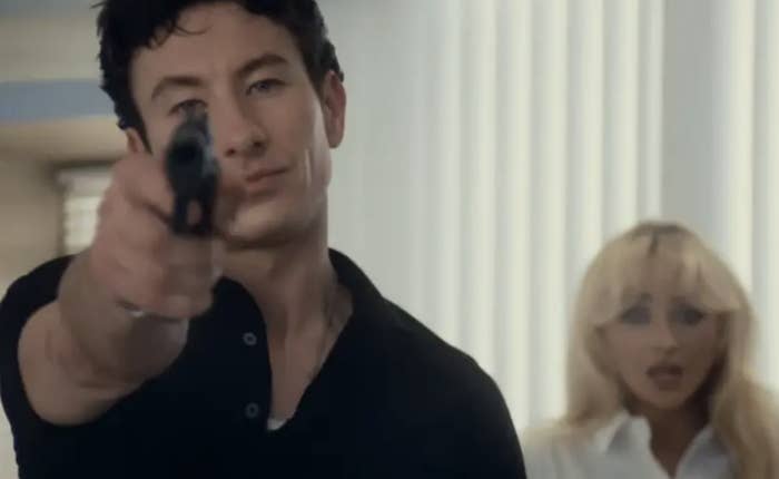 Barry Keoghan points a gun directly at the camera while Sabrina Carpenter looks scared in the background in a scene from the music video for “Please Please Please”