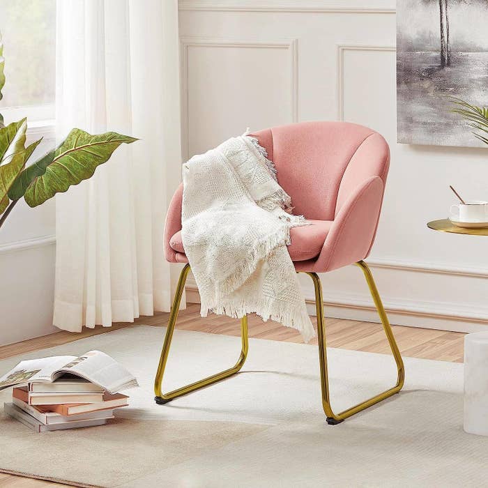 Stylish pink cushioned chair with gold legs, draped with a white throw blanket in a modern, elegant room with a side table and books