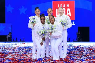 Simone Biles, Suni Lee, Jordan Chiles, Grace McCallum, and Jade Carey hold bouquets, posing together on a stage with confetti, in front of a "Team USA" background