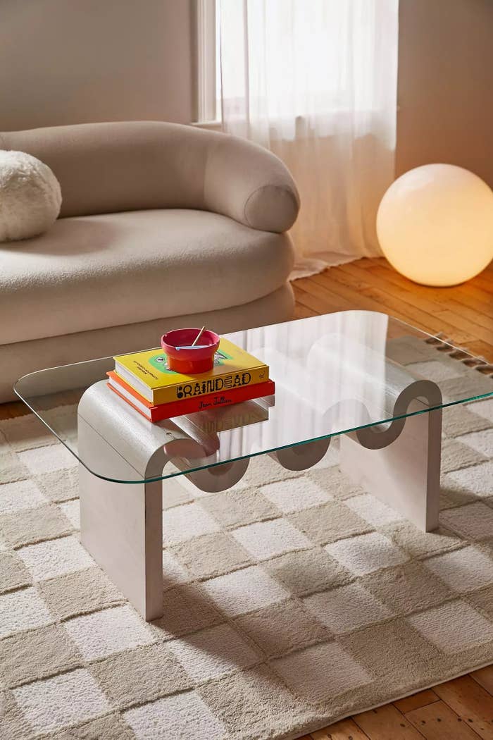 Modern living room setting with a unique glass coffee table. Books and a bowl are on the table. A sofa and a large round lamp are in the background