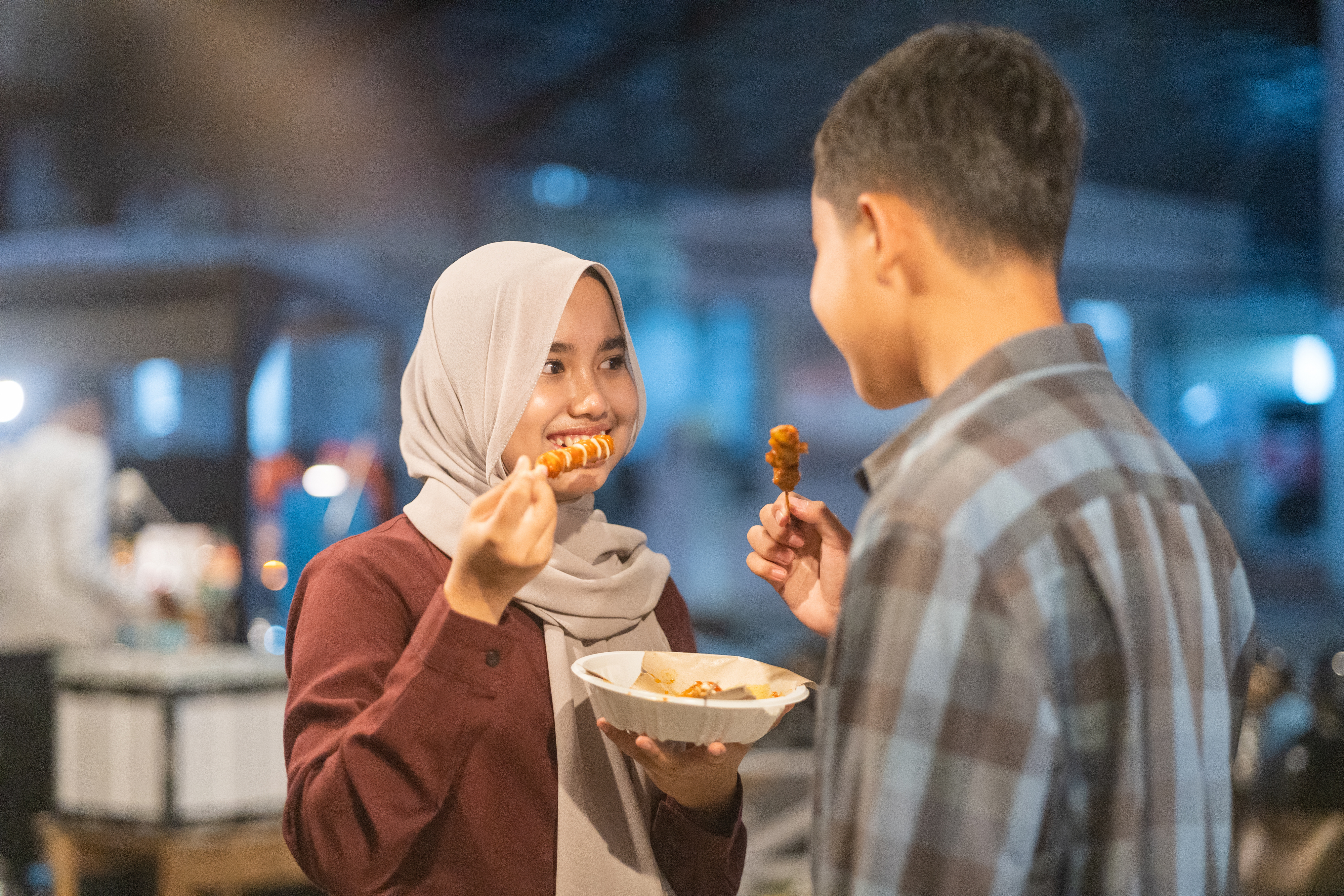 A woman and a man are sharing a meal outdoors. She is holding a skewer of food, smiling, and facing the man who is also holding a skewer