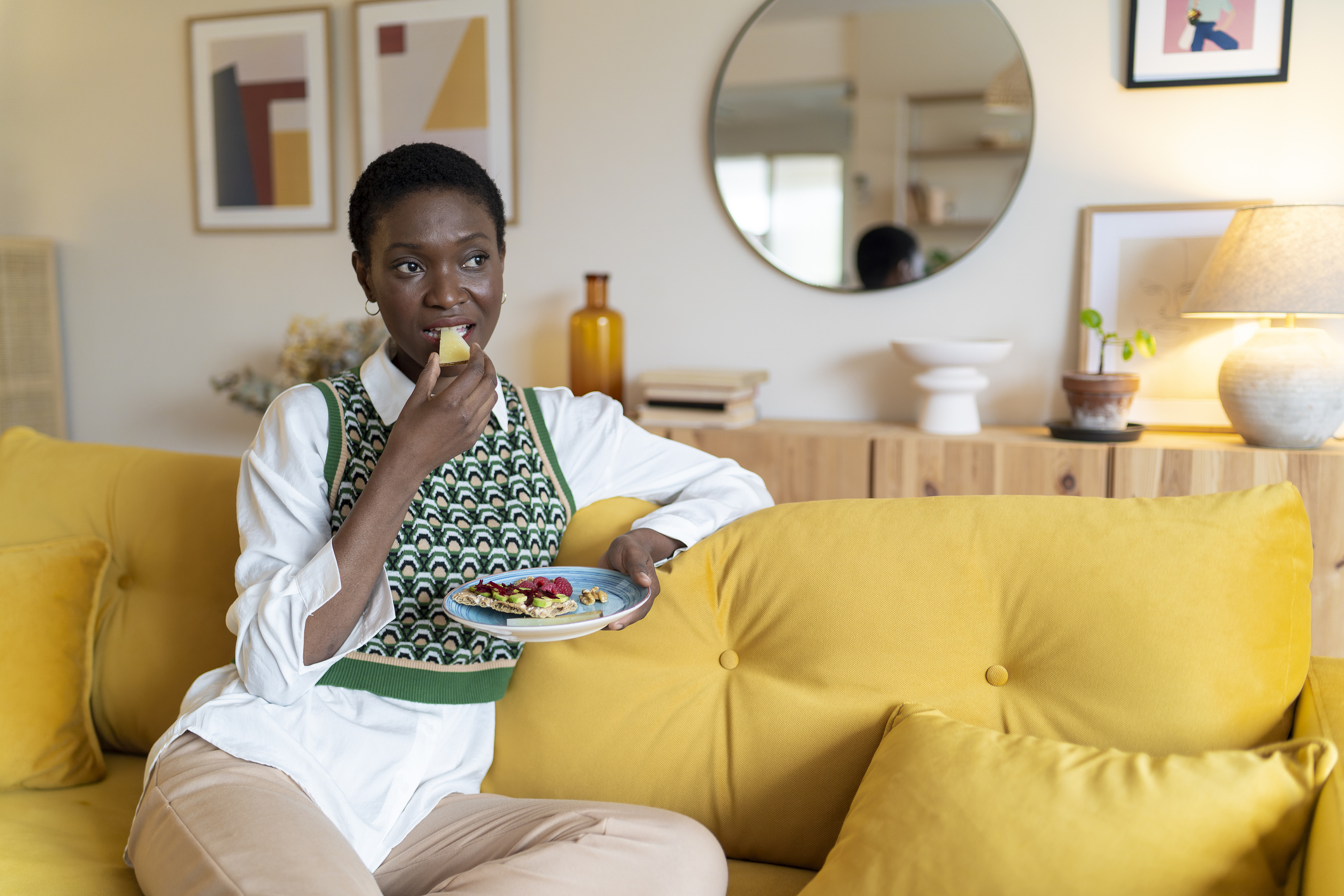 Person sits on a yellow sofa, enjoying a fruit platter in a modern, stylish living room with framed artwork and a round mirror in the background