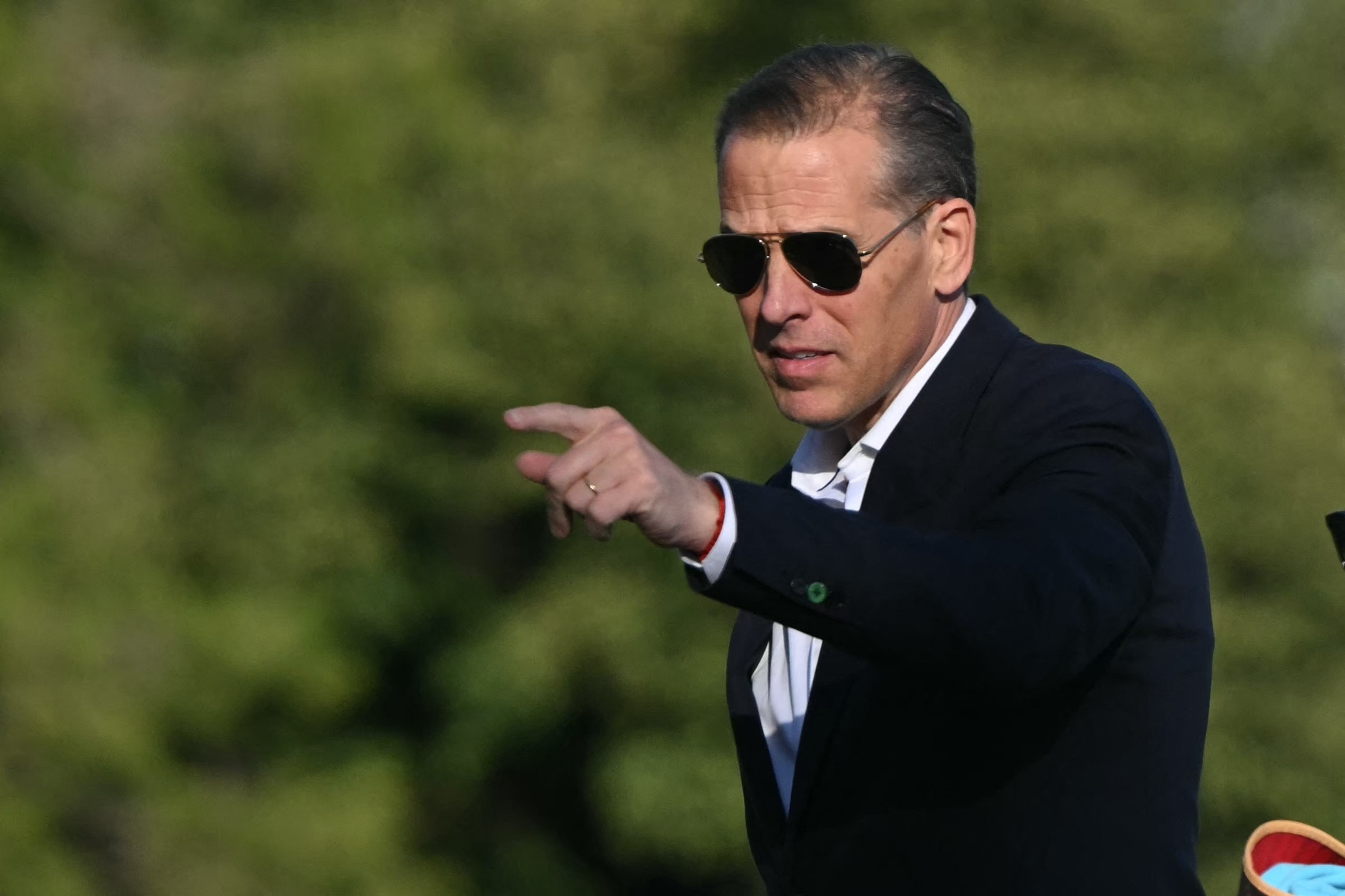 Hunter Biden is pointing, wearing a dark suit and sunglasses against a blurred outdoor background