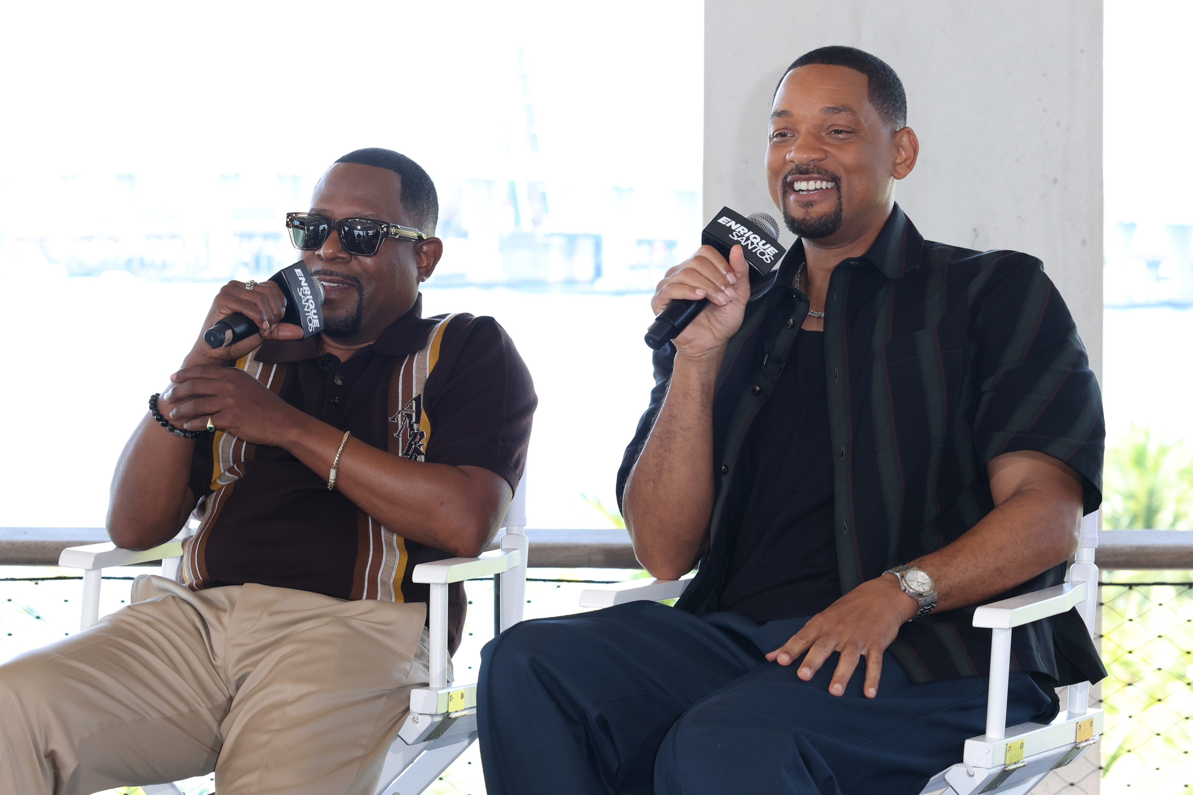 Martin Lawrence and Will Smith sit on director&#x27;s chairs, holding microphones and smiling during an interview