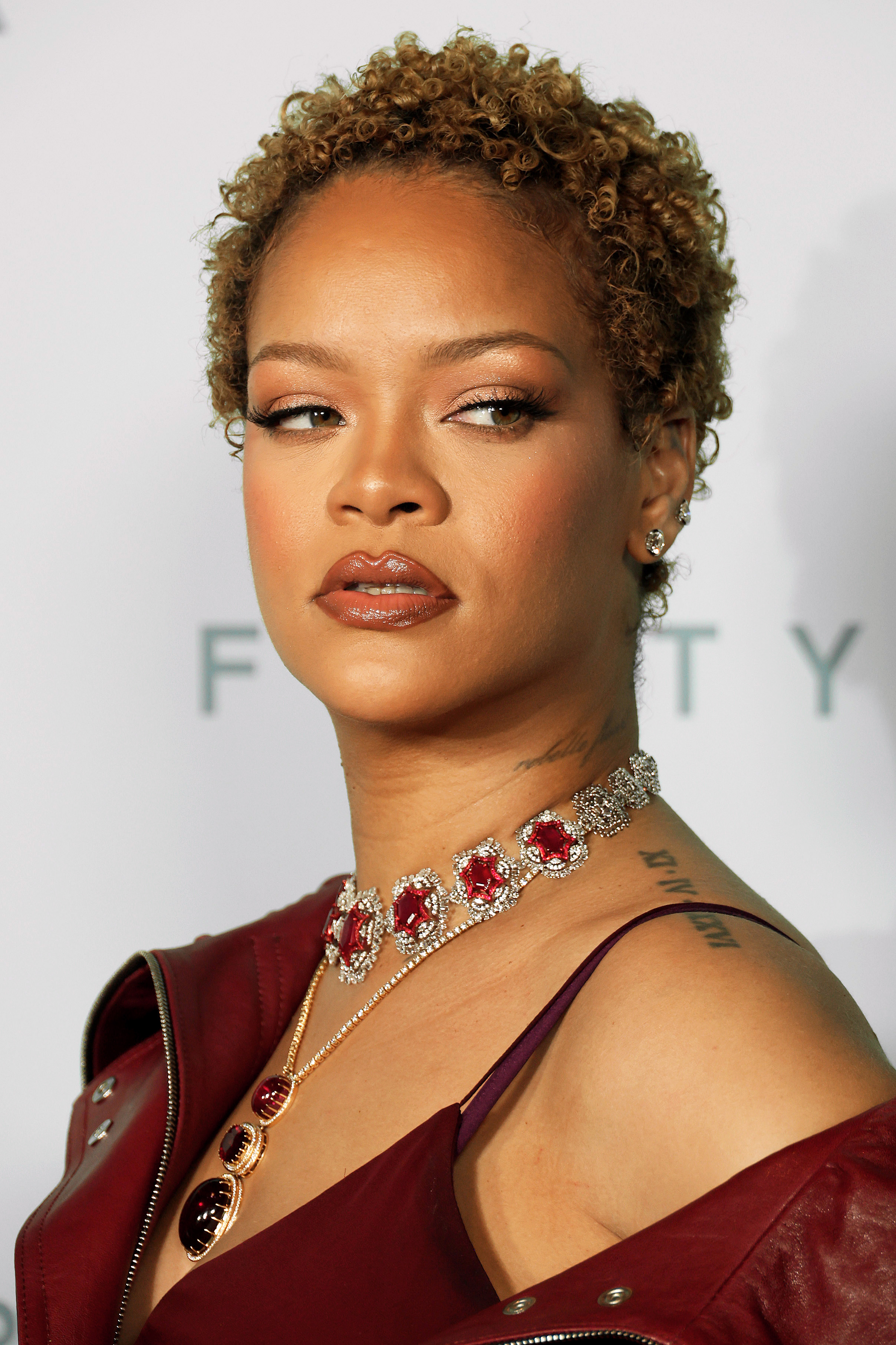 Rihanna, wearing an off-shoulder outfit, large red gemstone necklace, and matching pendant, looks to the side at a Fenty event