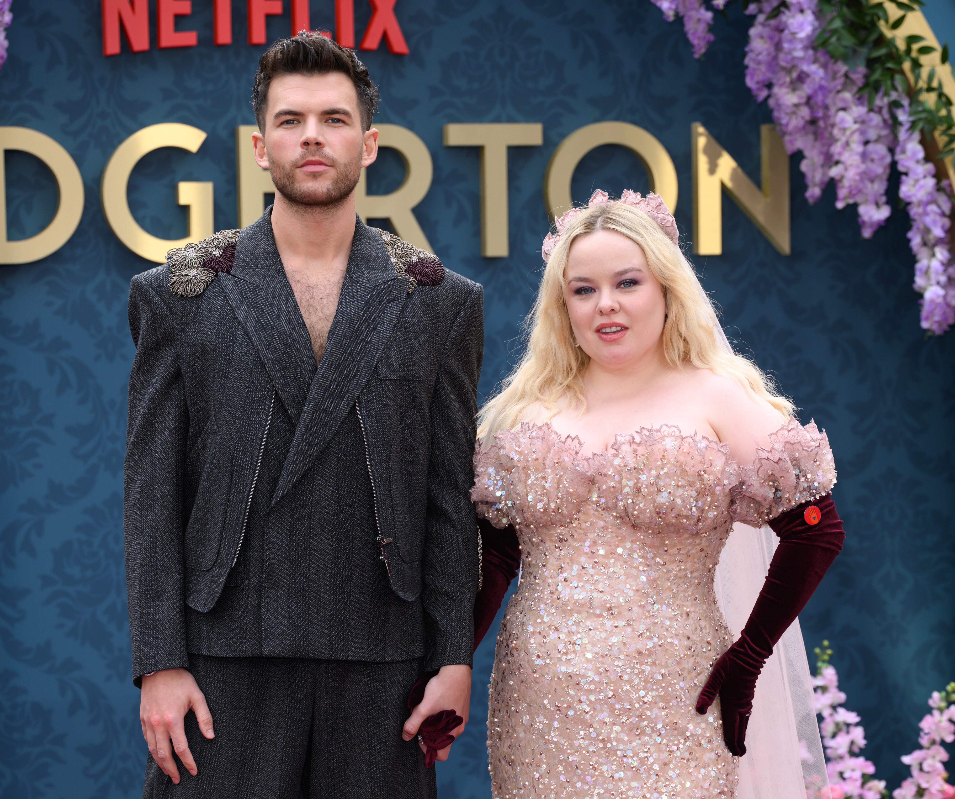 Luke Newton and Nicola Coughlan attend a Bridgerton event. Luke is in a stylish dark suit, and Nicola wears a sparkling off-the-shoulder dress with velvet gloves