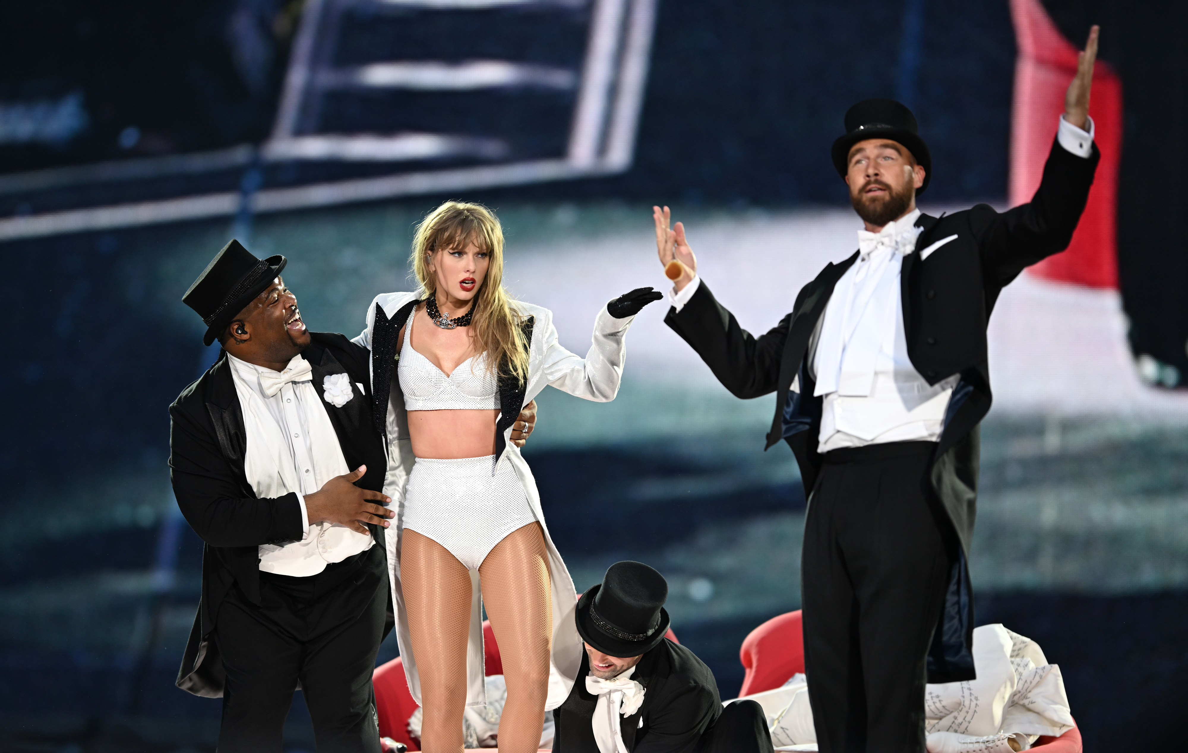 Taylor Swift performs on stage dressed in a white costume, accompanied by dancers in black tuxedos and top hats