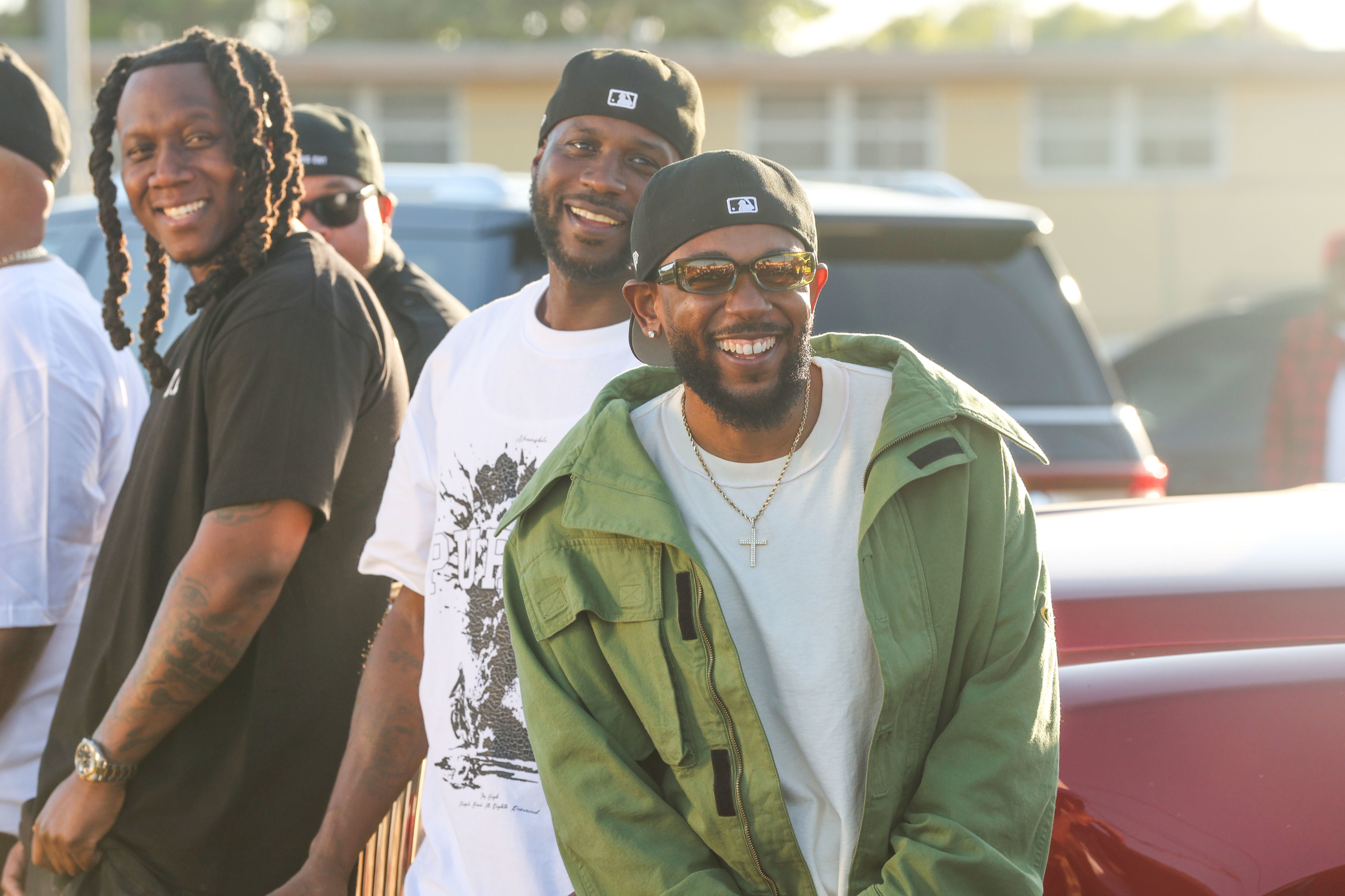 Three men, including Pusha T, smile and pose casually outdoors, wearing casual clothing and hats, in a cheerful and relaxed setting