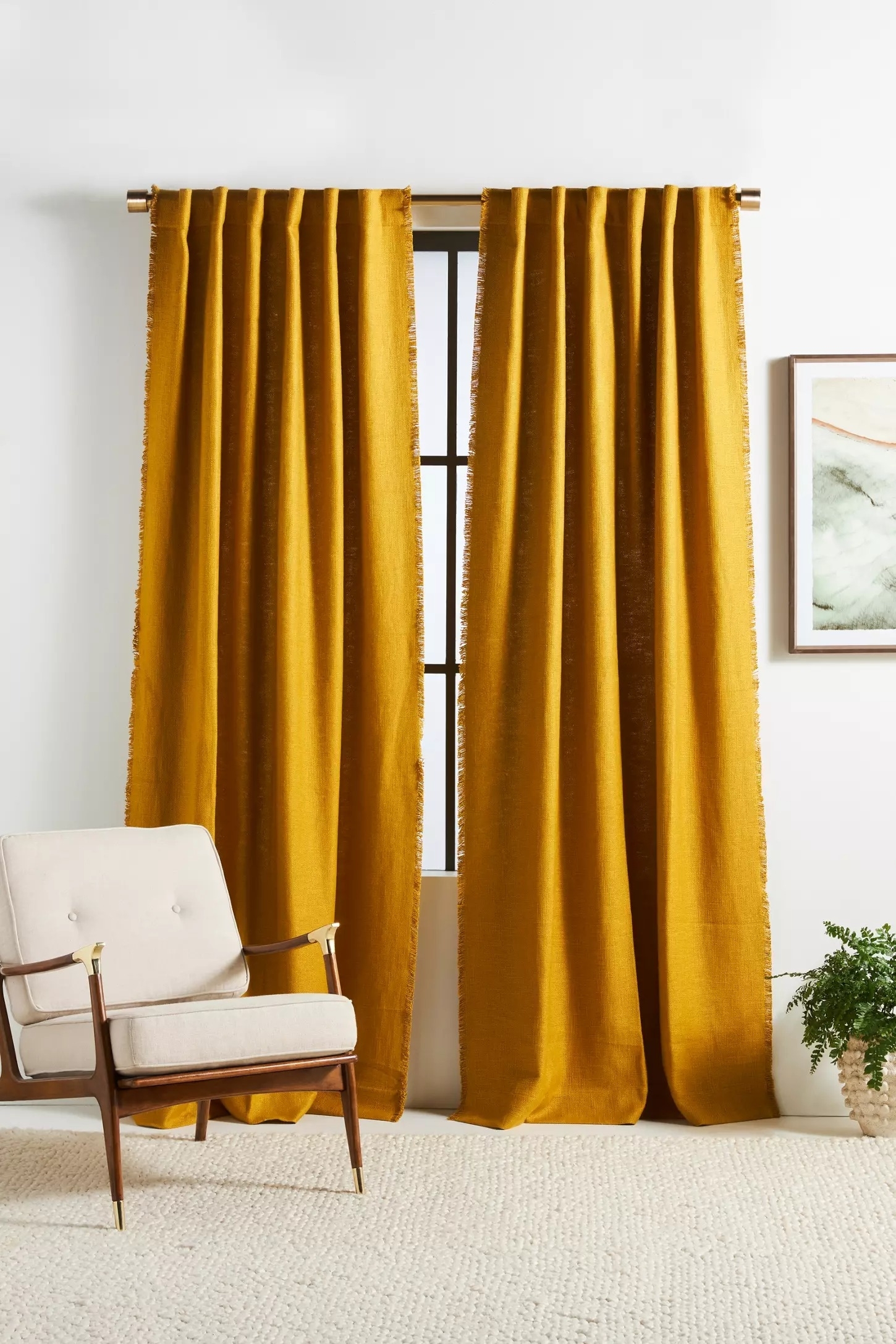 A stylish living room setup featuring mustard yellow drapes on a window, a beige mid-century modern armchair, a minimalist painting, and a potted plant