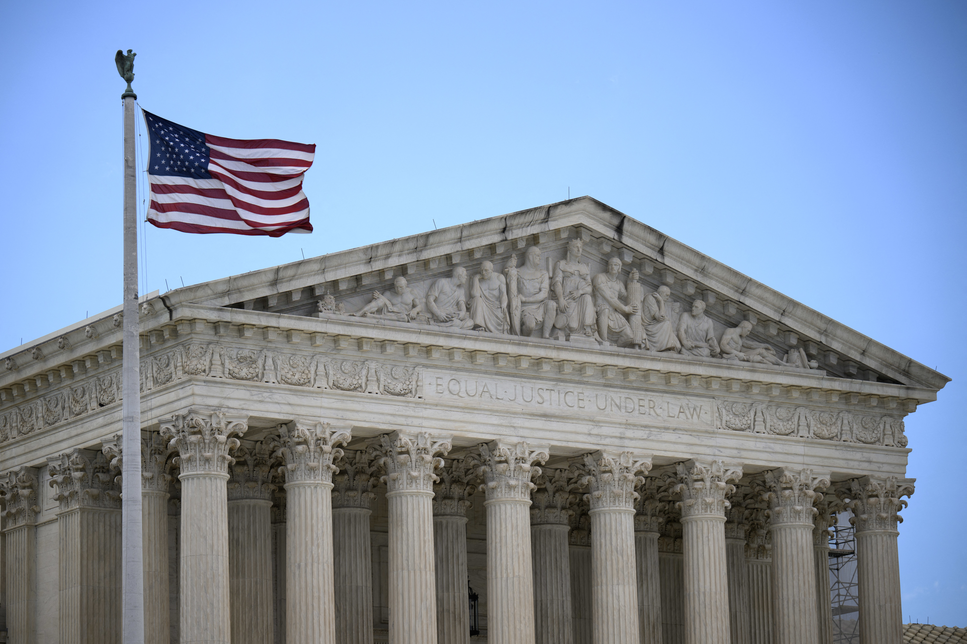 The image shows the front facade of the United States Supreme Court building with the American flag flying in the foreground. The inscription reads: &quot;EQUAL JUSTICE UNDER LAW.&quot;