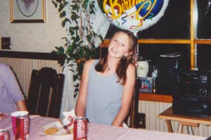 A young girl smiles while sitting at a table with a "Happy Birthday" balloon behind her. The table has food and soda cans. She is dressed casually