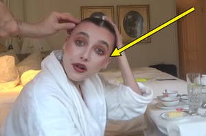 Emma Chamberlain, wearing a white robe in a hotel room, has makeup applied while she holds her shaved head. Arrow points to her, indicating focus on her