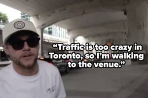 Niall Horan wears a cap and sunglasses, walking under an overpass with a line of cars in the background