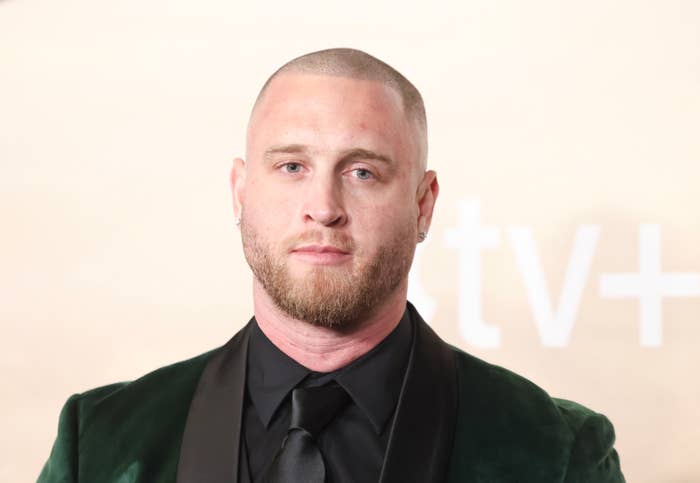 Chet Hanks with a shaved head and beard wearing a velvet suit jacket and dark shirt stands on the red carpet