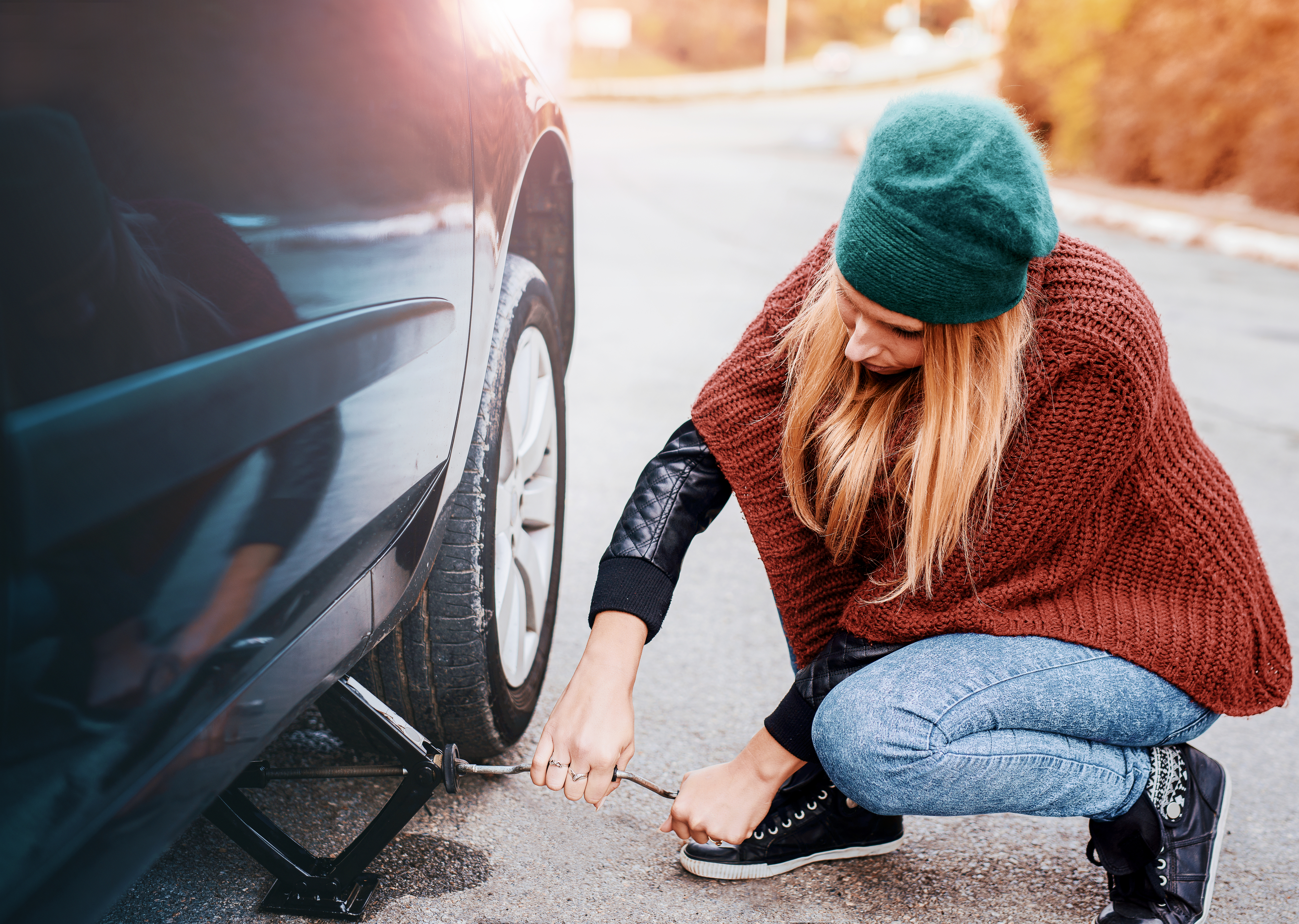 A woman with long hair, wearing a beanie, jeans, sneakers, and a poncho-style top, is changing a tire with a jack and wrench by the roadside